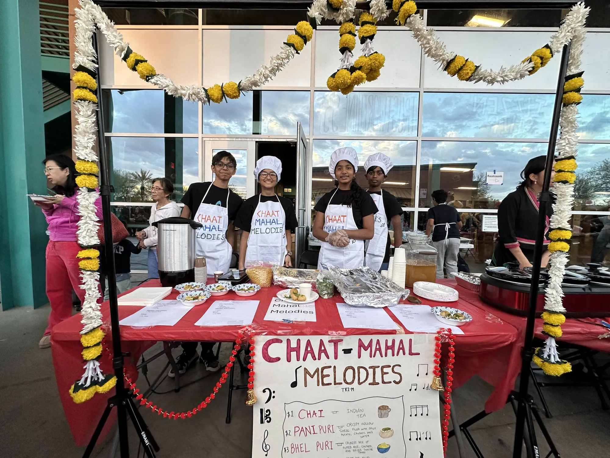 Students in chef hats serve chai and pani puri at a booth framed by marigold garlands