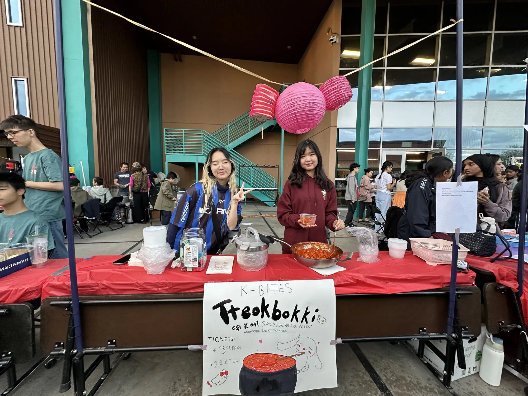 Students smile behind a tteokbokki booth with a hand-drawn sign